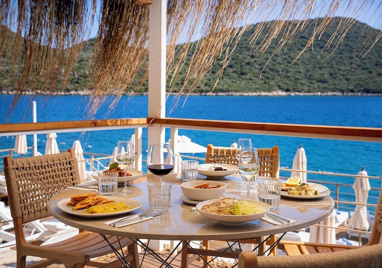 Une table à manger, dressée sur un balcon offrant une vue sur la mer et les collines, est garnie d’assiettes remplies de nourriture et de verres d’eau.