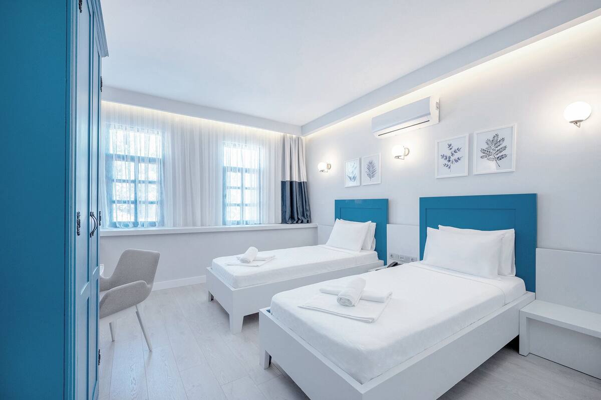 A hotel room with two beds, a desk, and a chair. There are windows with curtains, wall-mounted air conditioning, and framed artwork.
