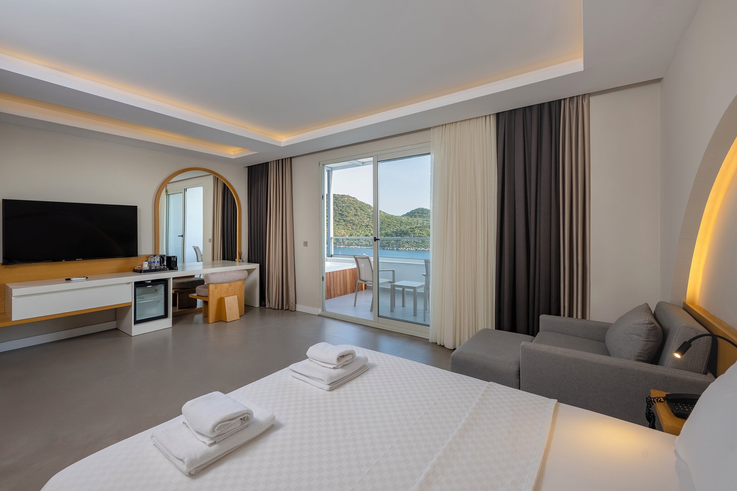 A modern hotel room with a large bed, a flat-screen TV, a desk with a chair, and a balcony with a view of the hills.