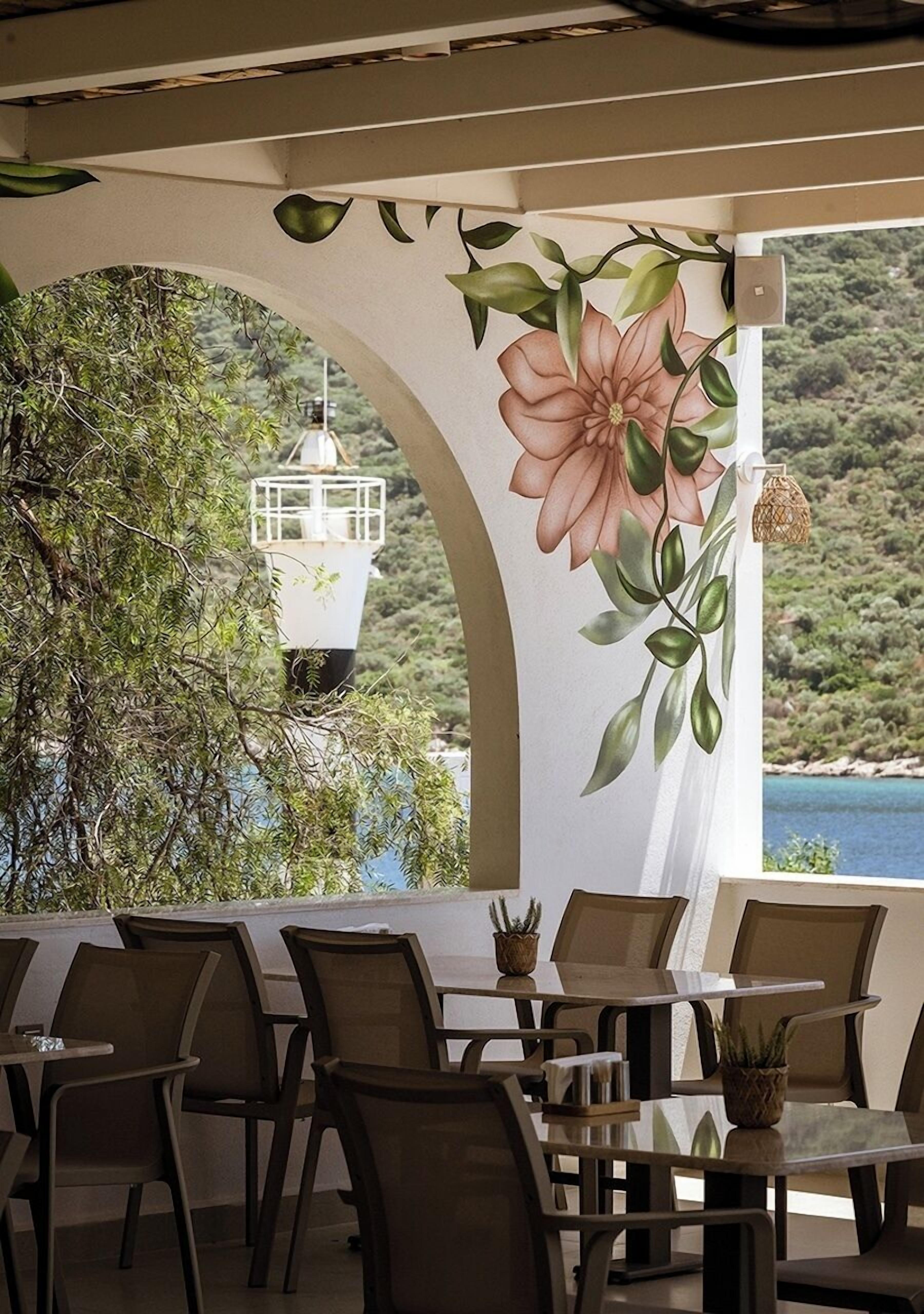 An outdoor seating area with tables and chairs, a large floral mural, and a view of the sea and hills.