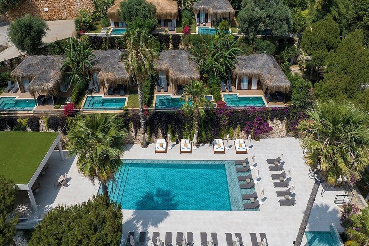 An aerial view of a resort with multiple thatched-roof structures, a central swimming pool, and lounge areas with sun loungers.
