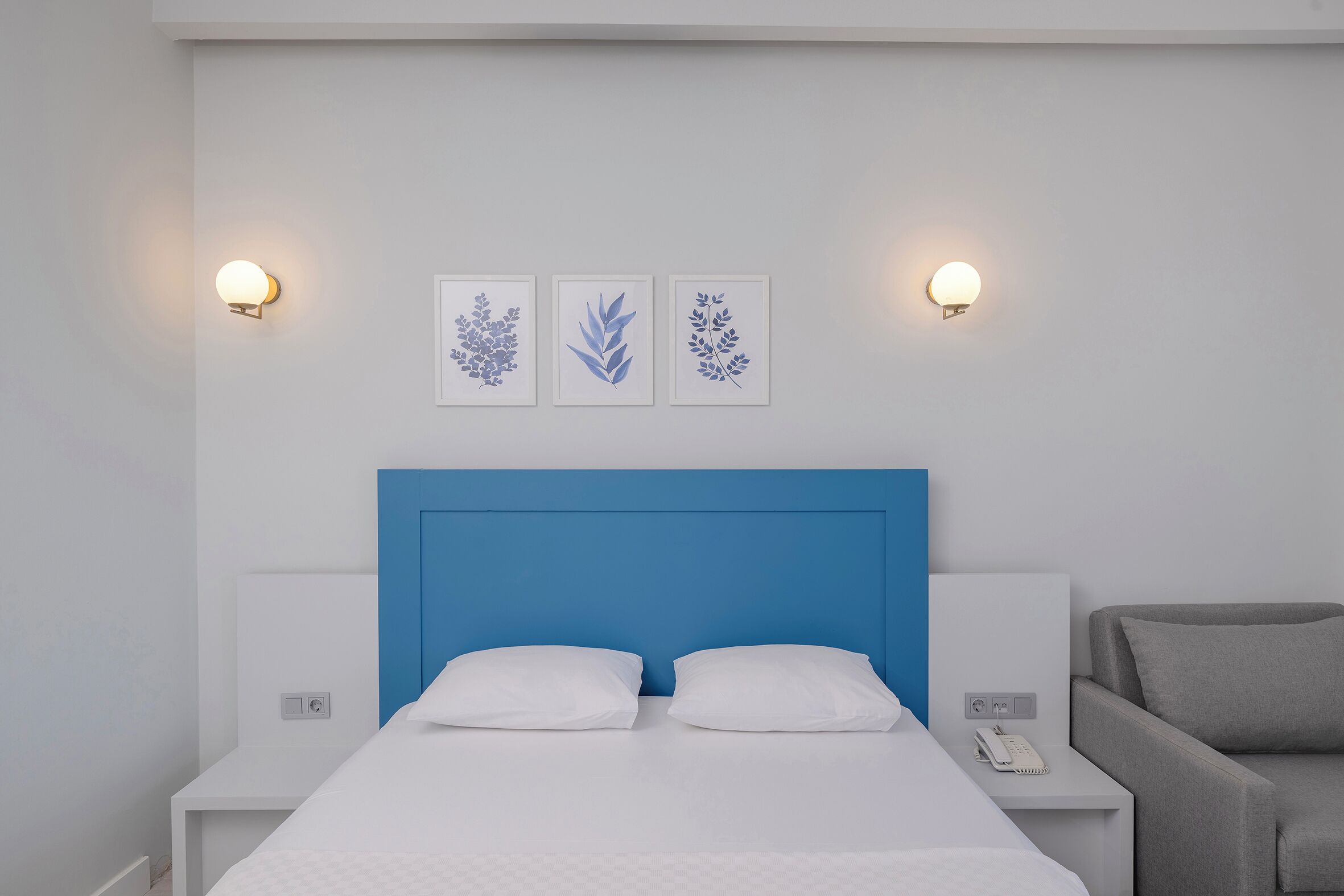 A modern hotel room with a blue headboard, white bedding, and wall-mounted light fixtures.