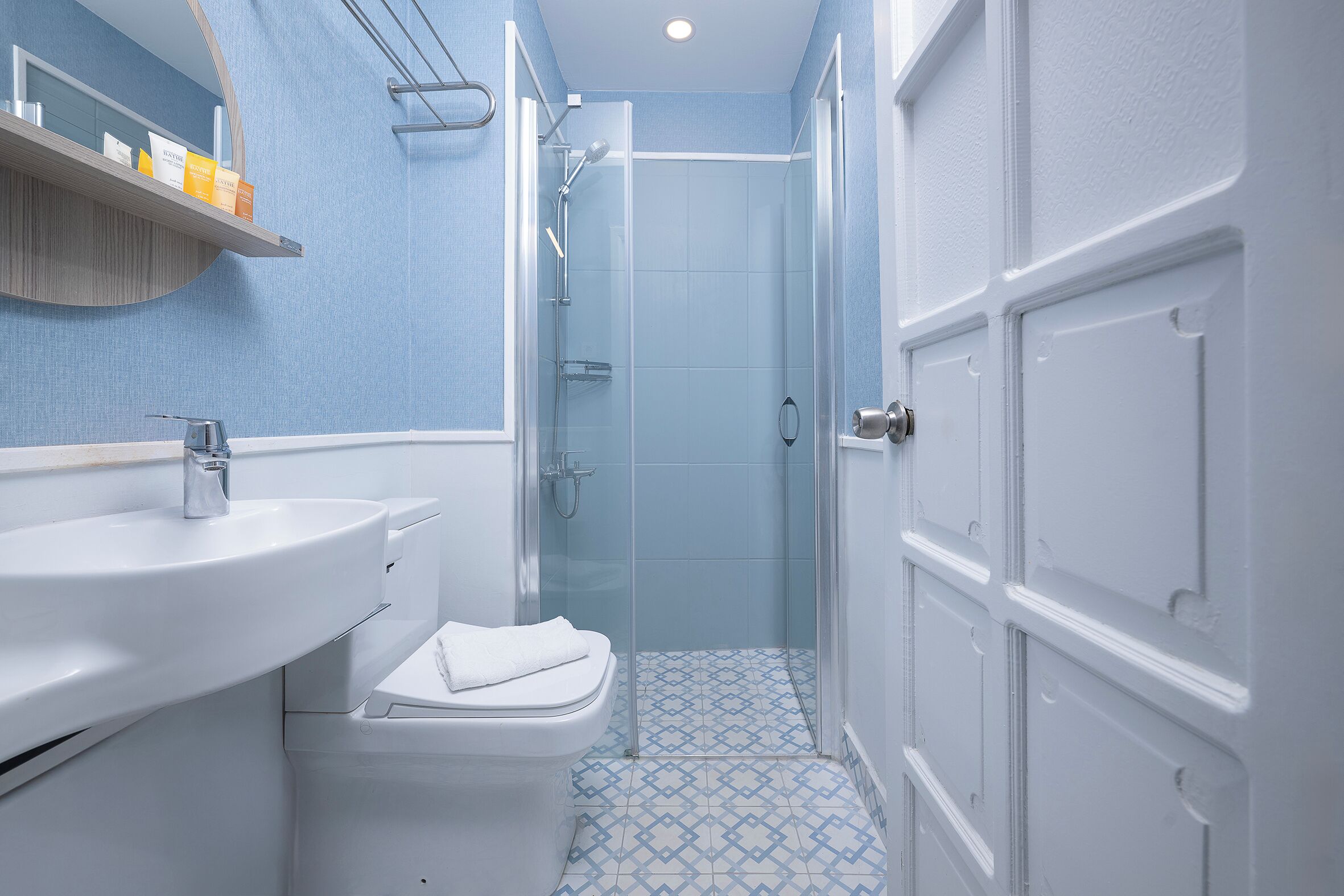 Une salle de bain avec un lavabo, des toilettes et une douche, le tout de couleur blanche. Une étagère contenant des articles de toilette se trouve au-dessus du lavabo.