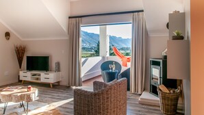 A modern living room with a large window, a flat-screen TV, a dining table, and a wicker chair. - Wind-Rose Guest House (Cape Town)