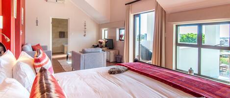 A hotel room with a large bed, a sitting area with a sofa and armchair, a TV, and a balcony with a view.