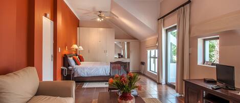 A spacious living area with a sofa, a coffee table with a vase, a TV on a wooden stand, and a bedroom with a bed and a desk.