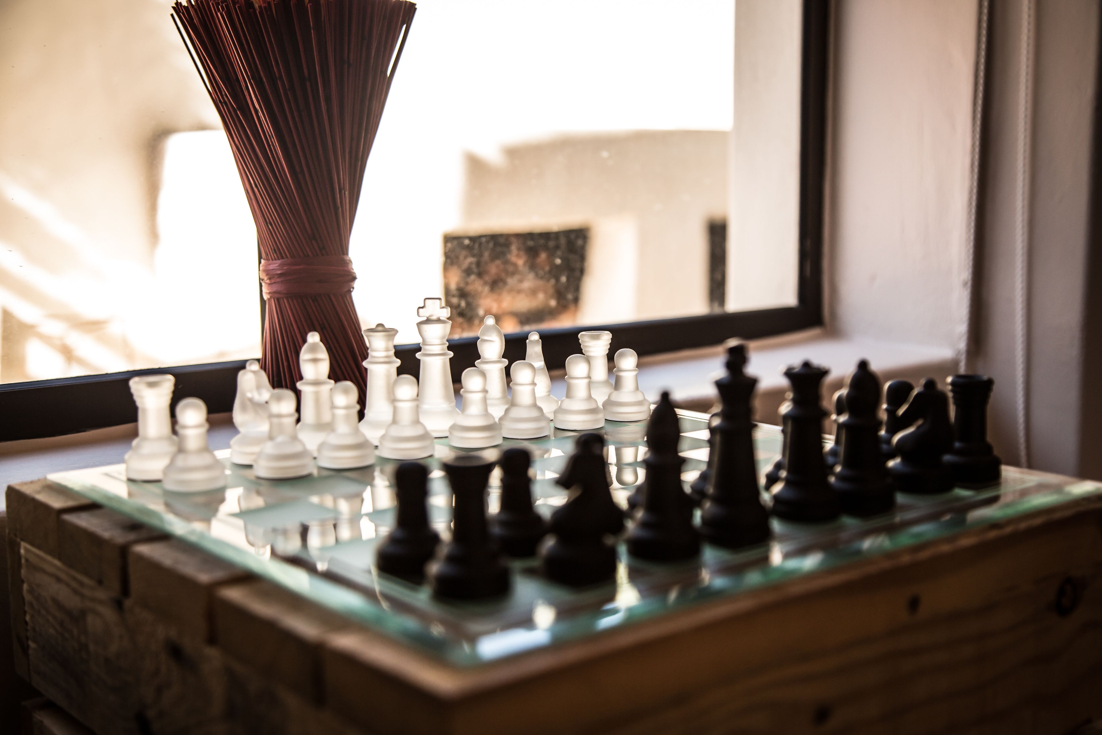 A chessboard with pieces on a glass table near a window.