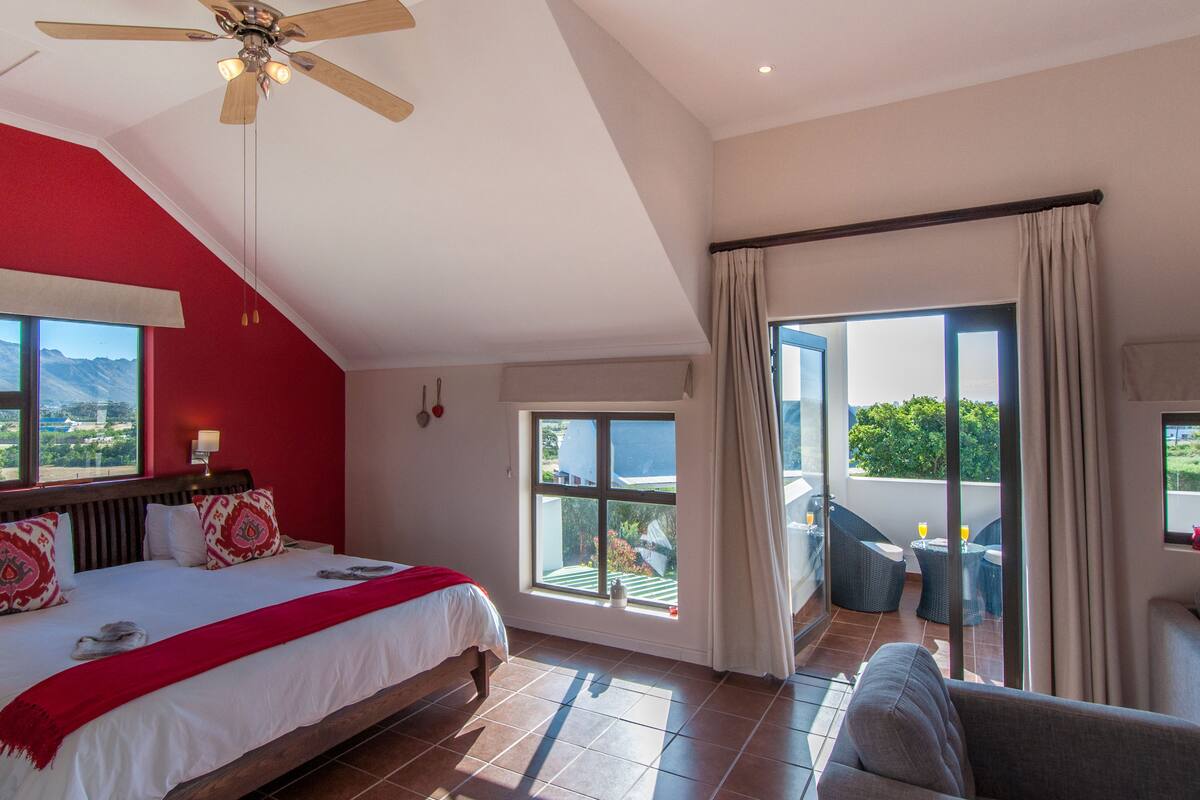 Une chambre dotée d’un grand lit, d’un ventilateur de plafond, d’une fenêtre avec des rideaux et d’un balcon aménagé avec des sièges.