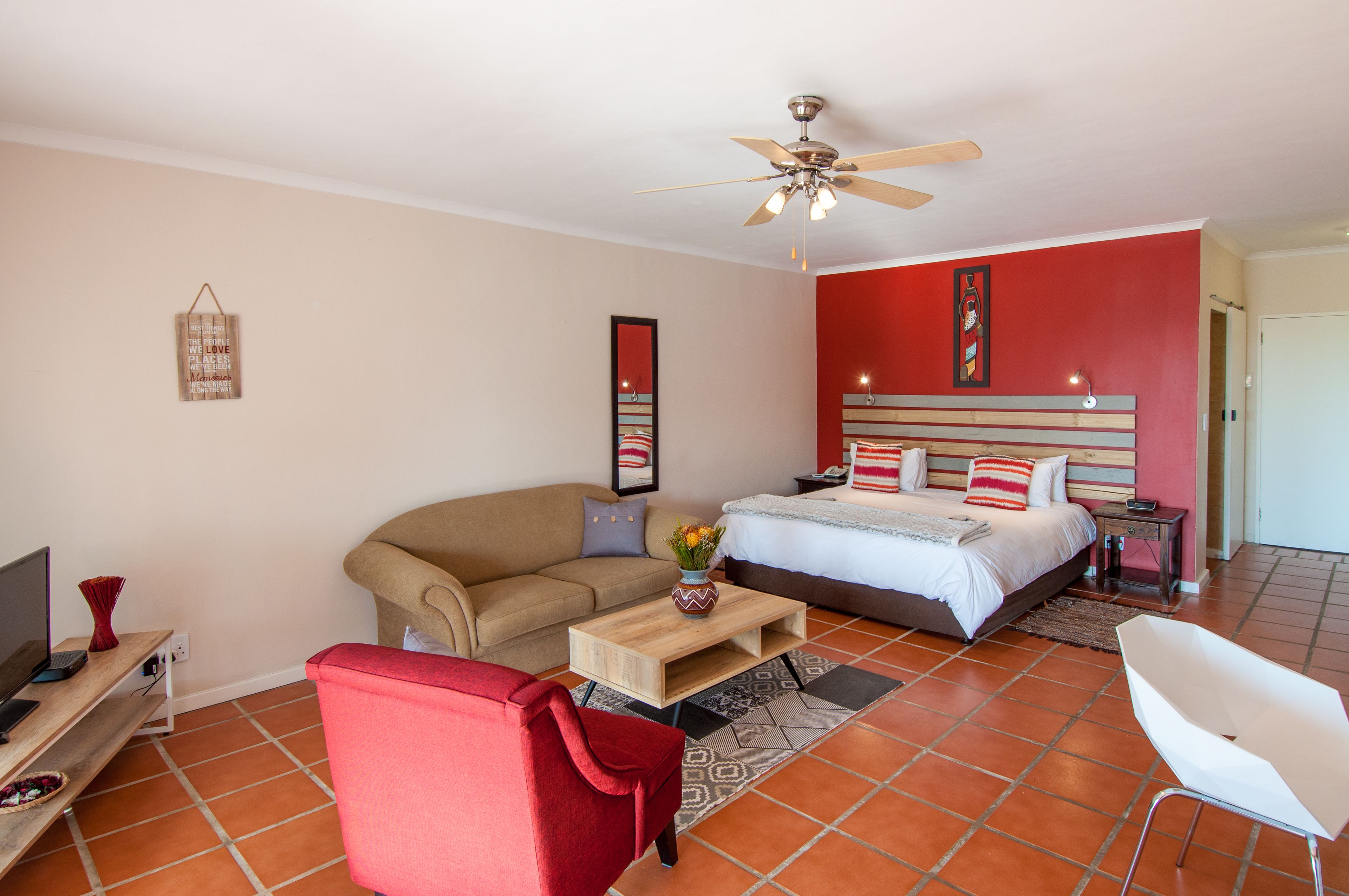 A modern bedroom with a bed, a sofa, a red armchair, a wooden coffee table, a ceiling fan, and a TV.