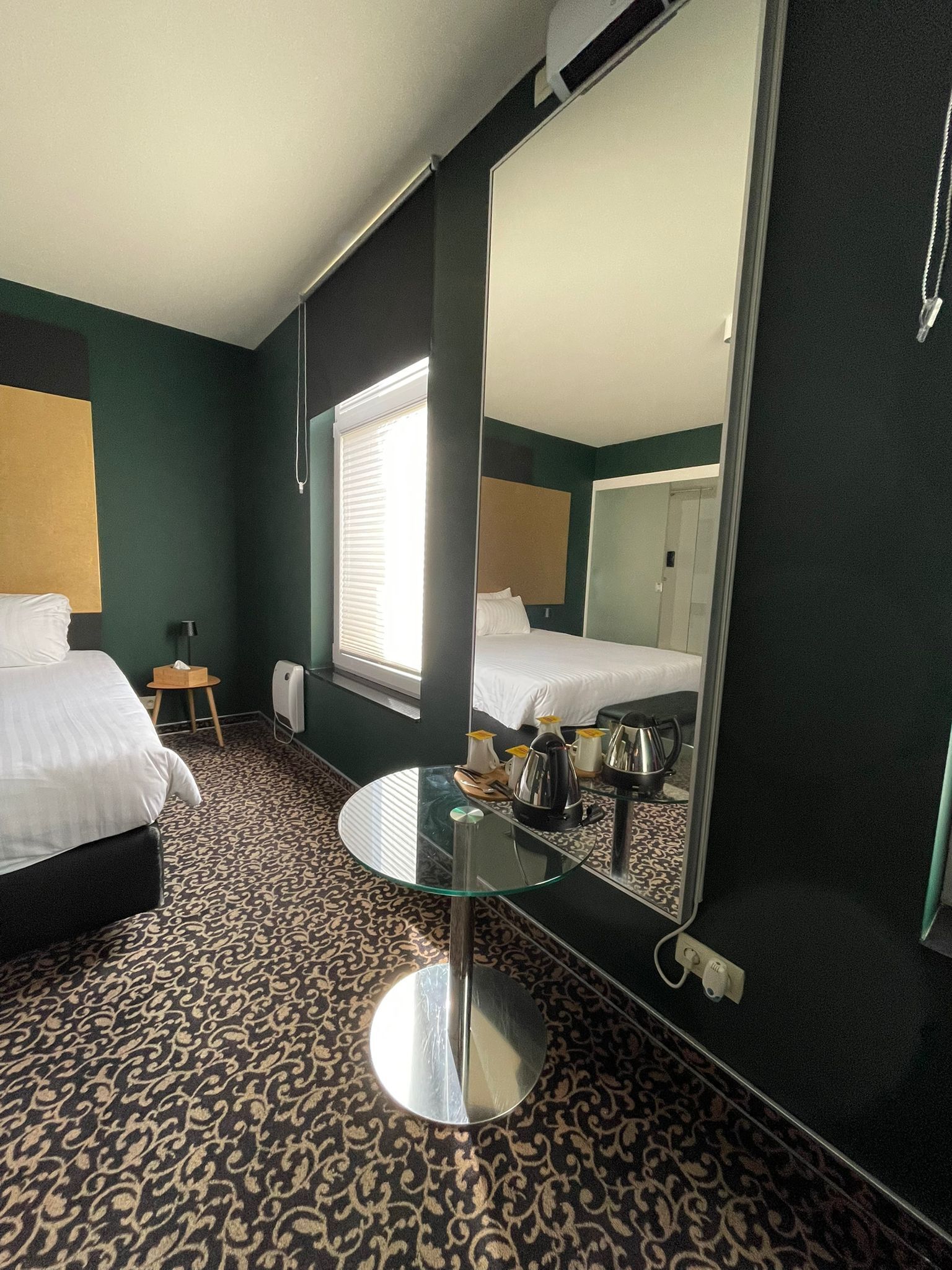 A hotel room with a bed, a round glass table, a mirror, and a window with blinds.