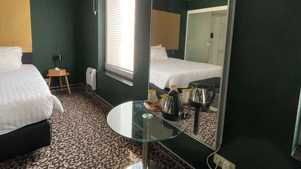 A hotel room with a bed, a round glass table, a mirror, and a window with blinds. - Tristar (La Louviere)