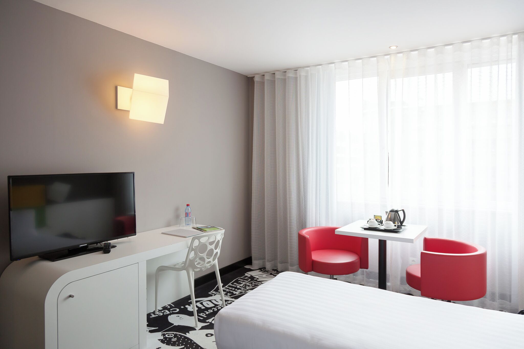 Una habitación de hotel con cama, televisión, escritorio con silla, una mesita con dos sillas rojas y ventana con cortinas translúcidas.