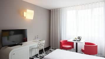 Una habitación de hotel con cama, televisión, escritorio con silla, una mesita con dos sillas rojas y ventana con cortinas translúcidas.