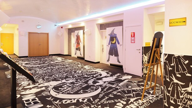A hotel hallway with a patterned carpet, doors with artwork, a fire alarm, and a sign for Room 101. - Tristar (La Louviere)