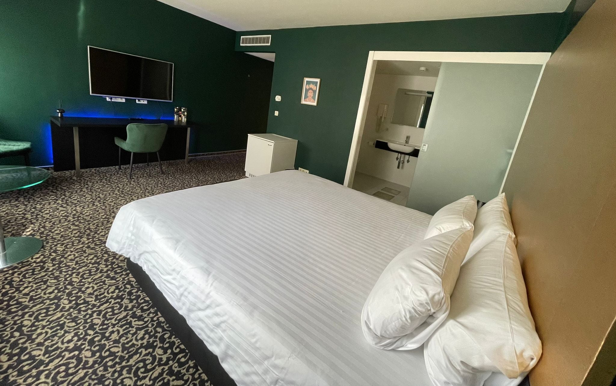 Une chambre d’hôtel avec un grand lit, un bureau avec une chaise, un téléviseur à écran plat et une salle de bain visible à travers une porte ouverte.