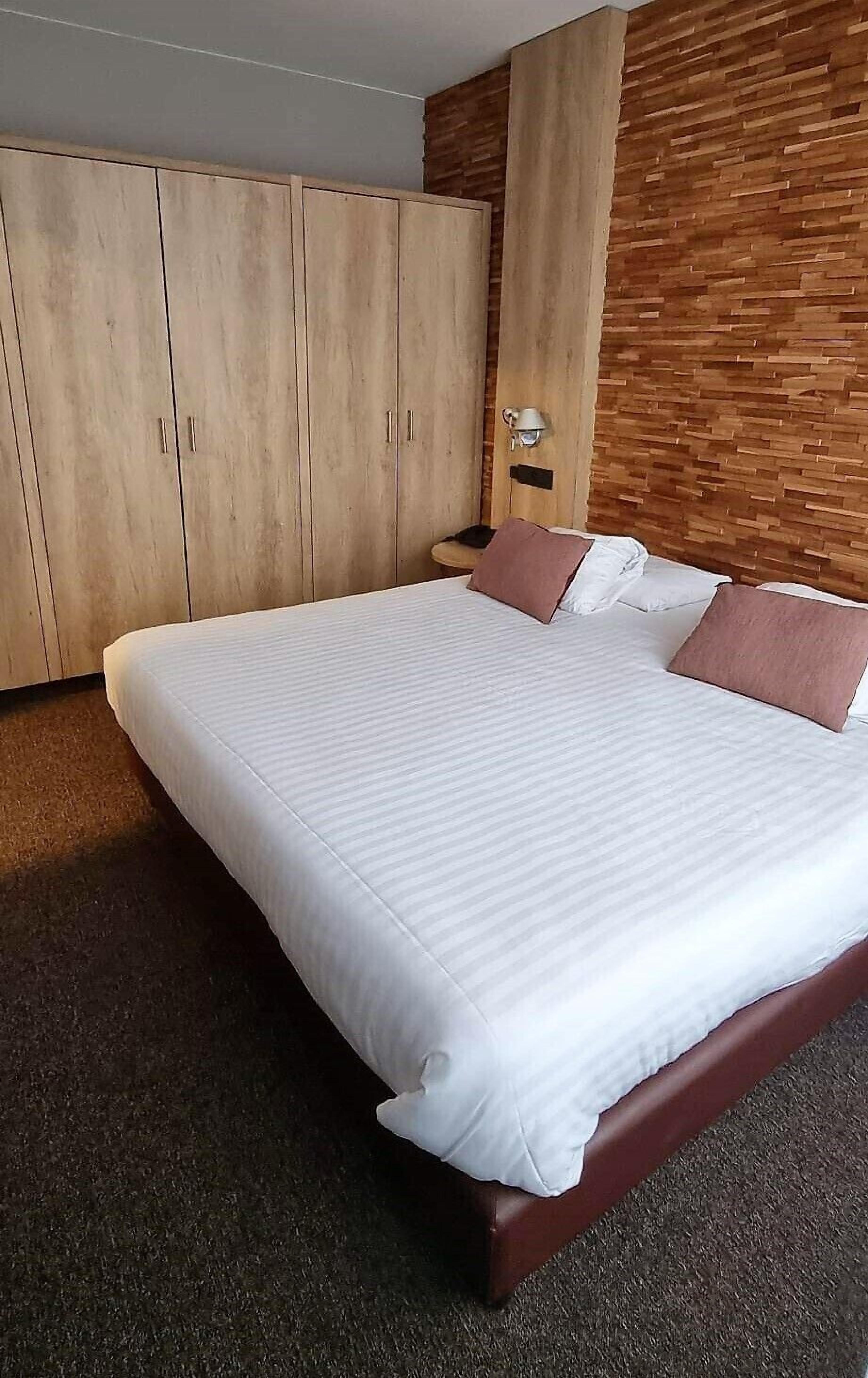 Un lit bien fait, avec une literie blanche et deux coussins roses, dans une pièce dotée d’armoires en bois et d’un mur à motifs.