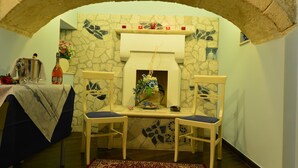 A room with a stone fireplace, two chairs, a table with a tablecloth, a vase with flowers, and a rug.