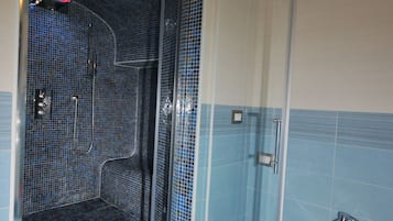 Egy fürdőszoba, melyben zuhanyzó található, a falakat kék mozaikkák borítják, egy fehér mosdókagyló és egy üveg zuhanyajtó szolgálja a kényelmet.