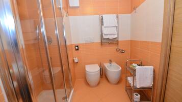 A bathroom with a glass-enclosed shower, a toilet, a bidet, and a shelf with towels.