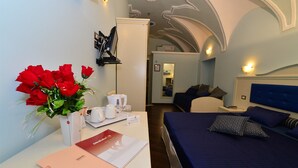 A hotel room with a bed, a desk with a vase of red roses, a coffee maker, and a red booklet.