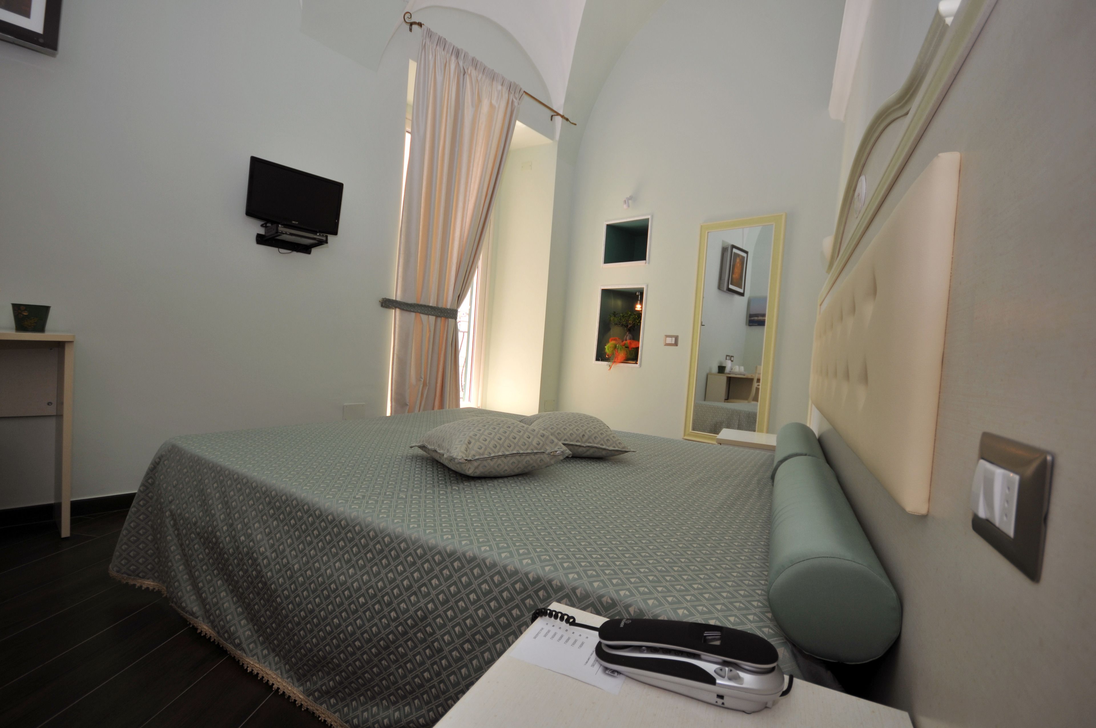 A bedroom with a bed, a bedside table with a book and a phone, a TV mounted on the wall, a window with curtains, and a mirror on the wall.