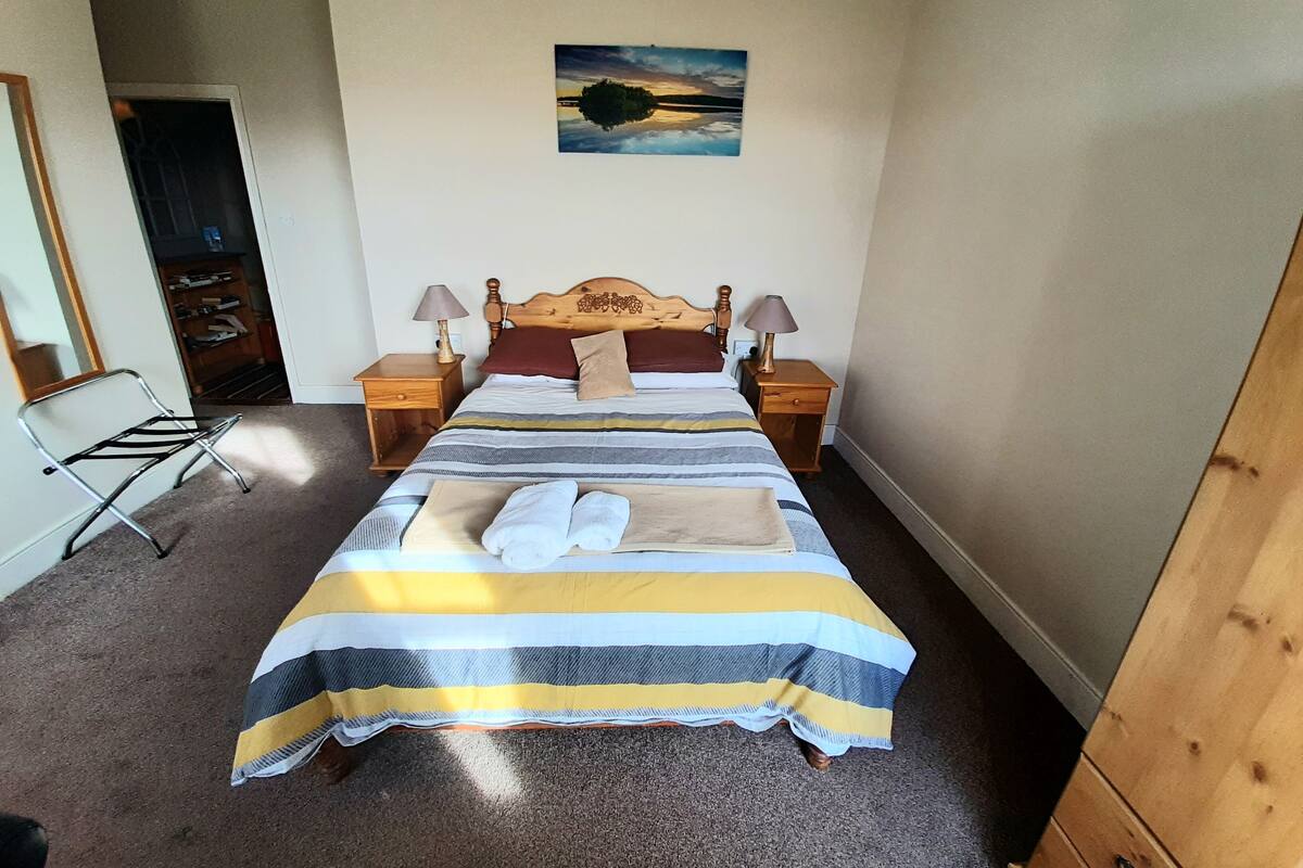 A neatly made bed with striped bedding, two bedside tables with lamps, a wooden headboard, and a painting of a sunset on the wall.