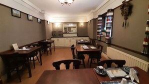 Un comedor con mesas y sillas de madera, una araña y una cocina al fondo.