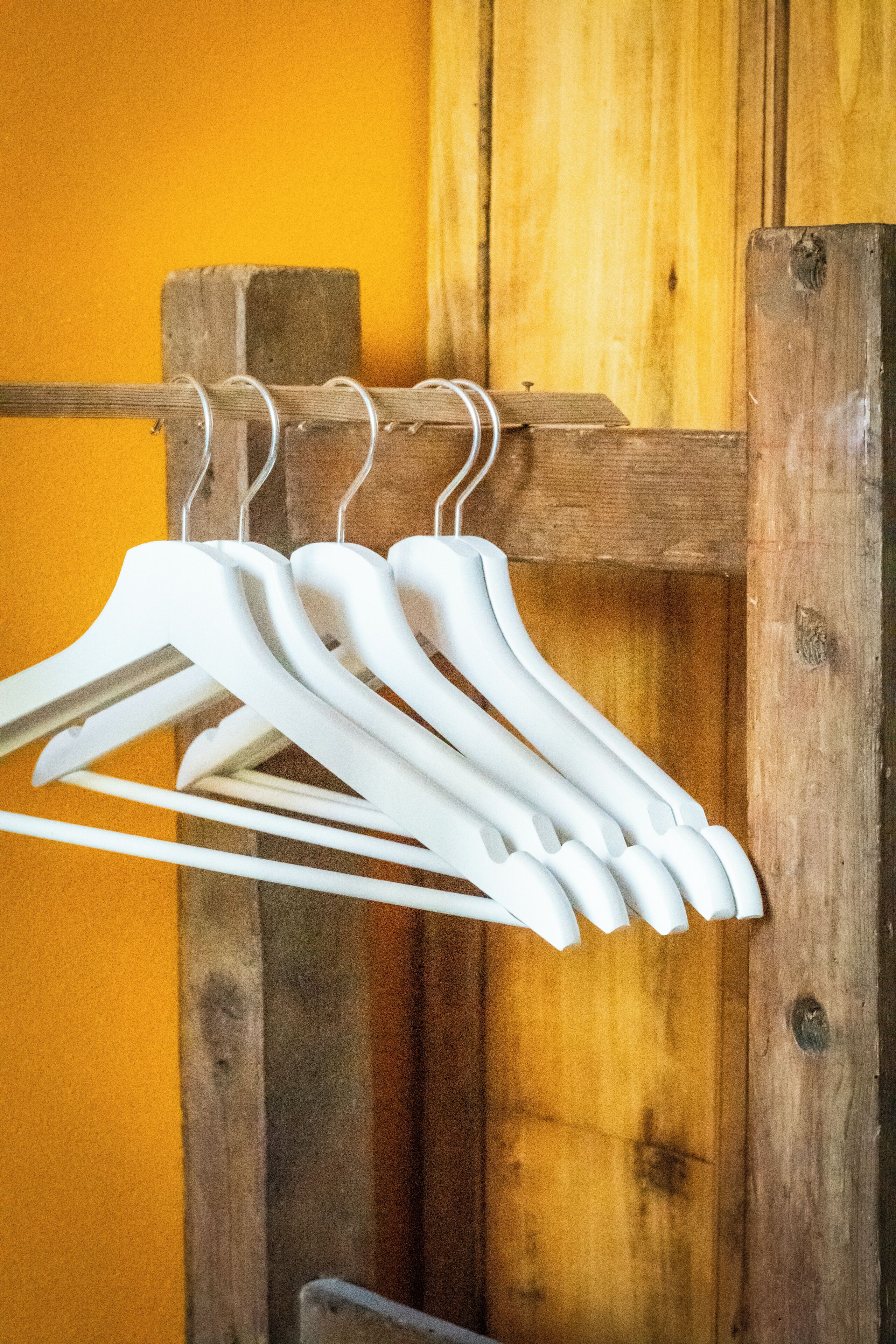 A set of white hangers hanging on a wooden hanger rod against a yellow wall.