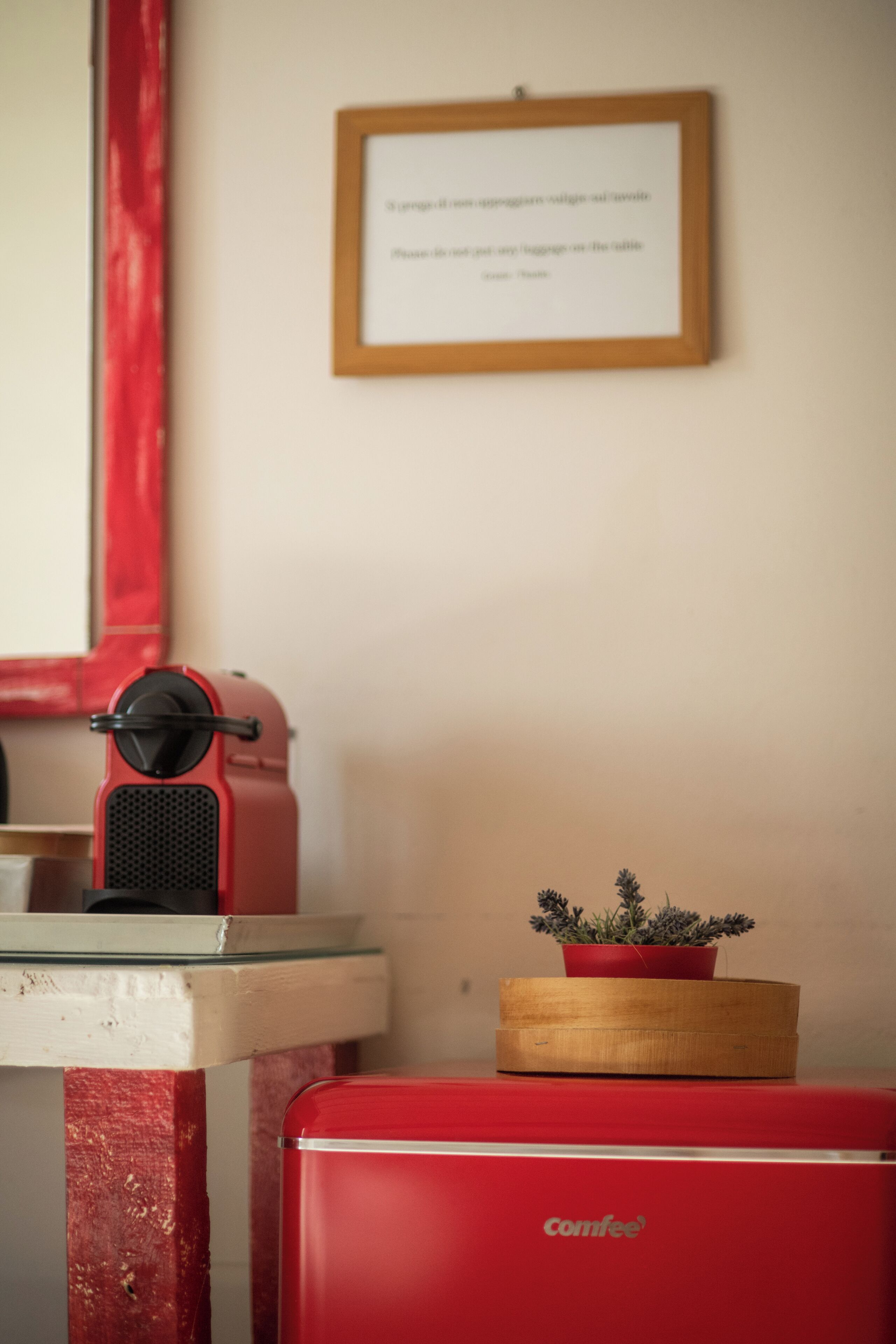 A red vintage-style coffee machine on a table, a framed notice on the wall, and a small potted plant on a red cabinet.