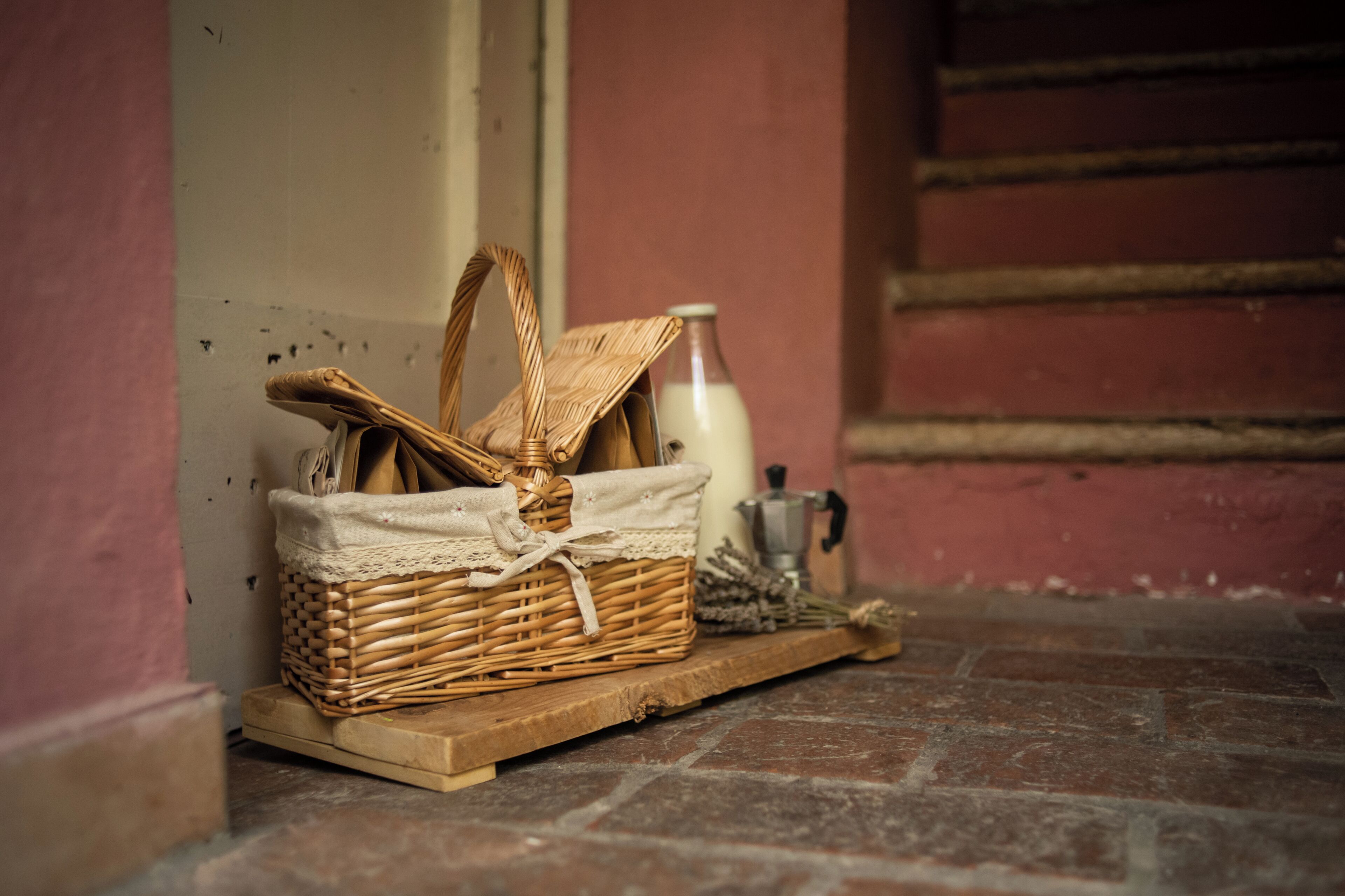A wicker basket with a cloth cover on a wooden platform, a milk bottle, and a coffee pot on a brick floor.