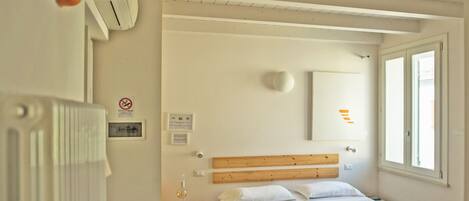Habitación de hotel con cabecera de madera, cama con ropa de cama blanca, mesita de noche con lámpara y ventana con persianas.