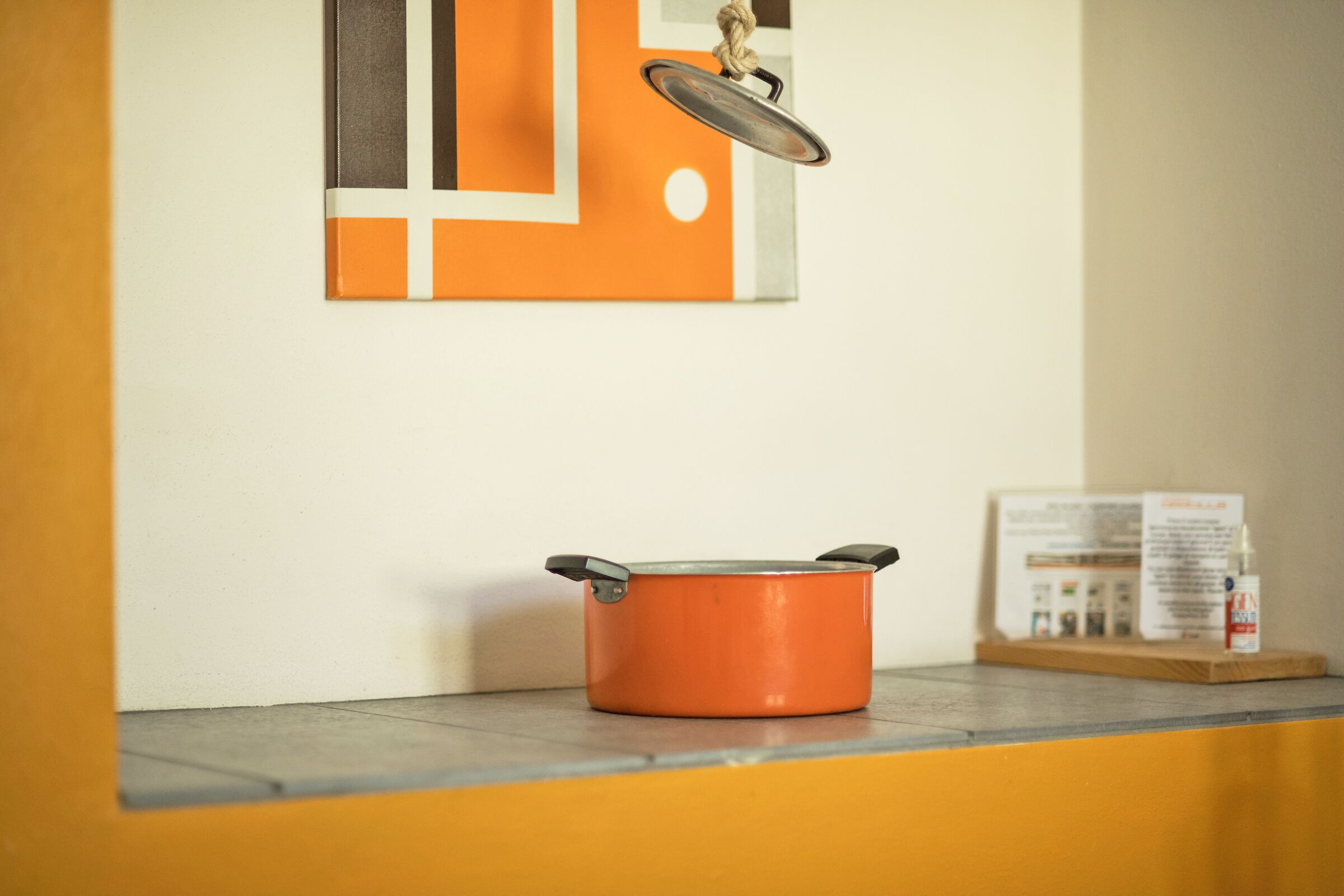 A kitchen with an orange pot on the counter, a hanging pot lid, and a framed picture on the wall.