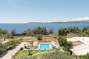 Zona de piscina con vistas al mar, rodeada de vegetación y algunos edificios.