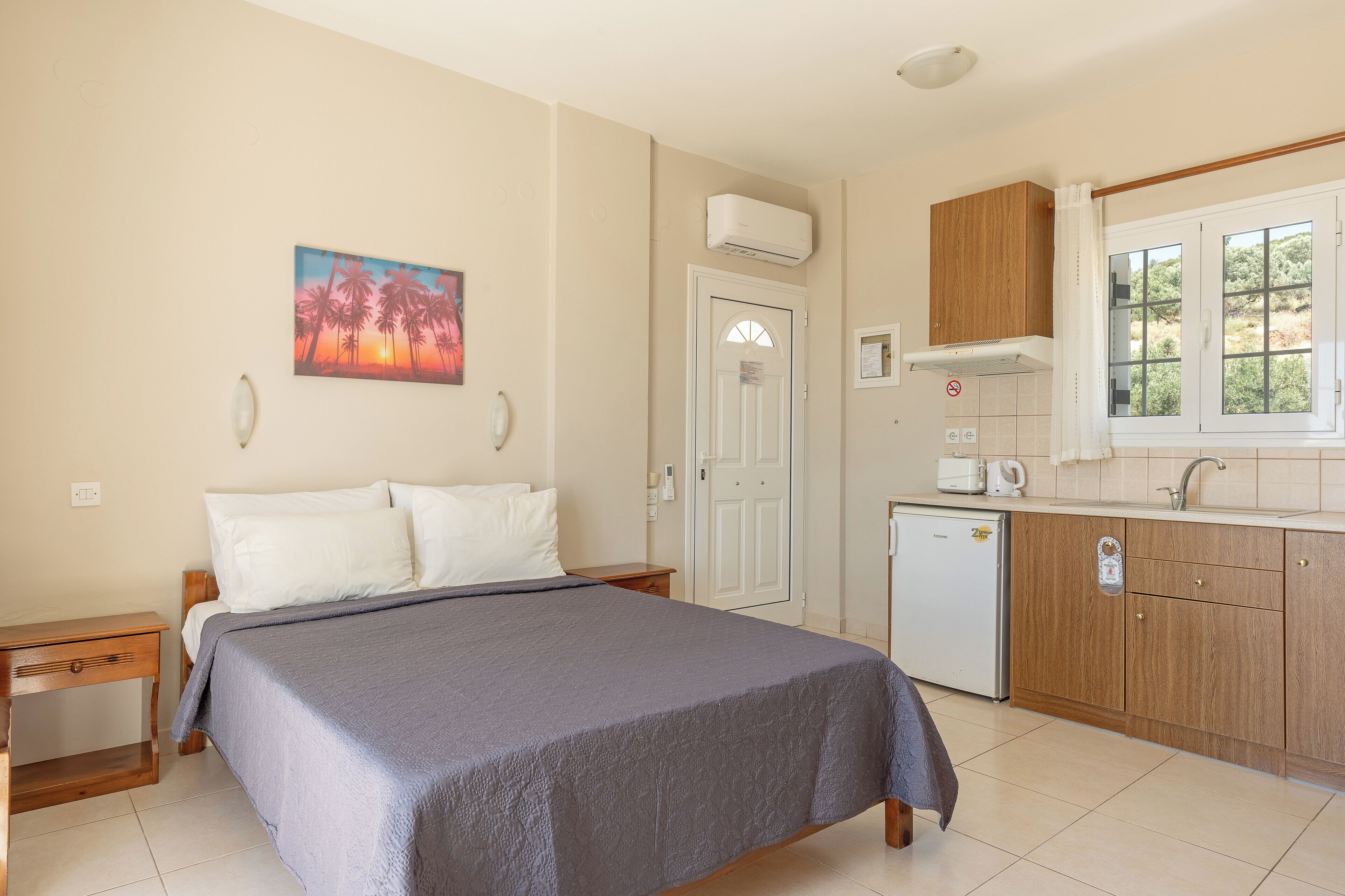 A bedroom with a bed, a nightstand, a painting, a door, a kitchen area with cabinets, a sink, and a window.