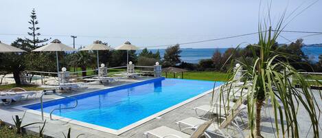 A swimming pool with lounge chairs and umbrellas, surrounded by greenery and a view of the sea.