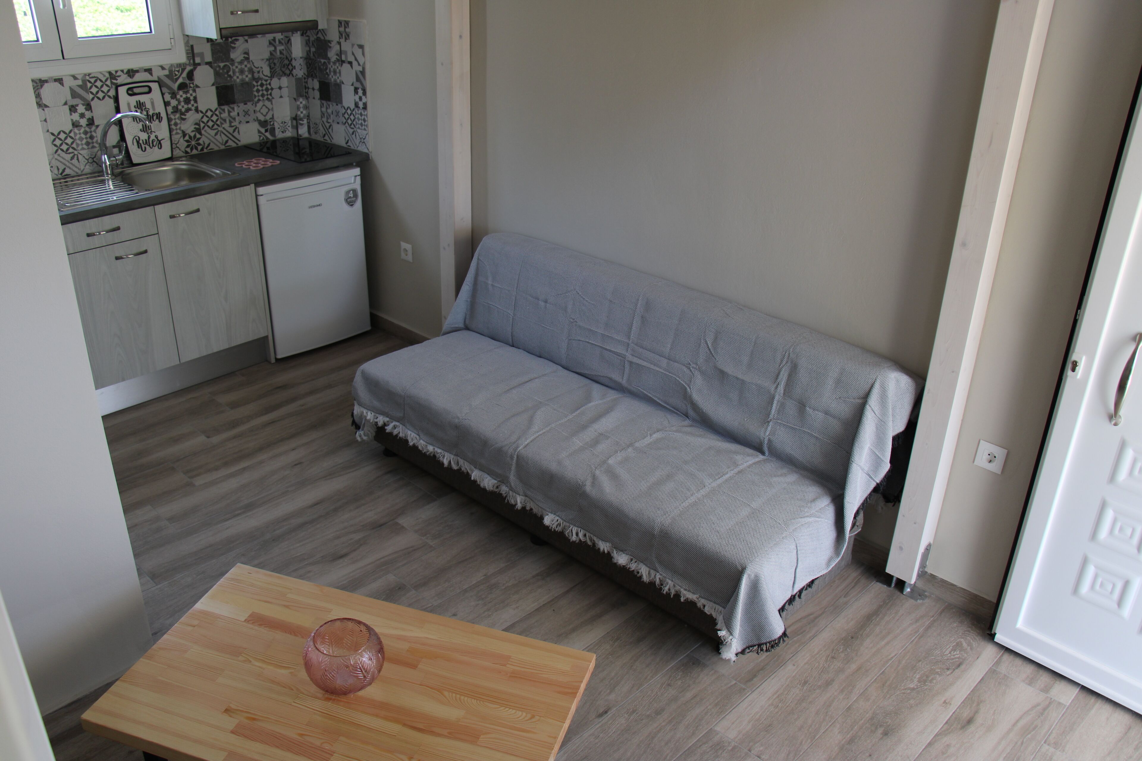 A compact kitchen with a sink, dishwasher, and a small table with a glass object. A grey sofa is positioned against the wall.