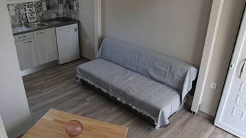 A compact kitchen with a sink, dishwasher, and a small table with a glass object. A grey sofa is positioned against the wall.
