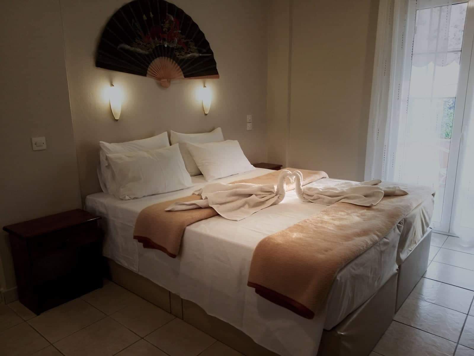 A neatly made bed with a decorative fan and wall-mounted lights, a bedside table, and a view of the outdoors through a window.