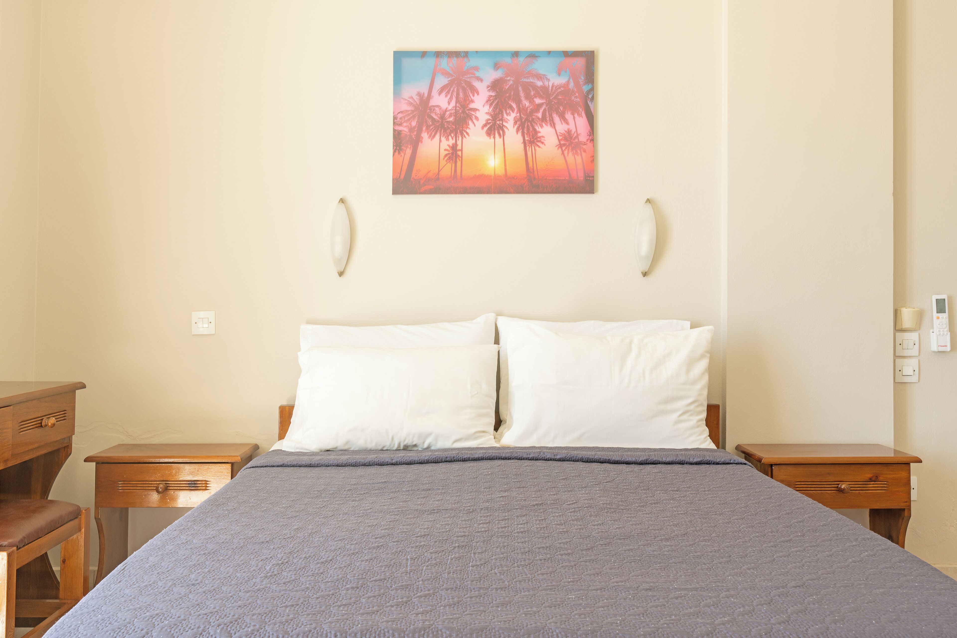 A bedroom with a bed, two nightstands, a wooden desk, and a painting of palm trees and a sunset on the wall.