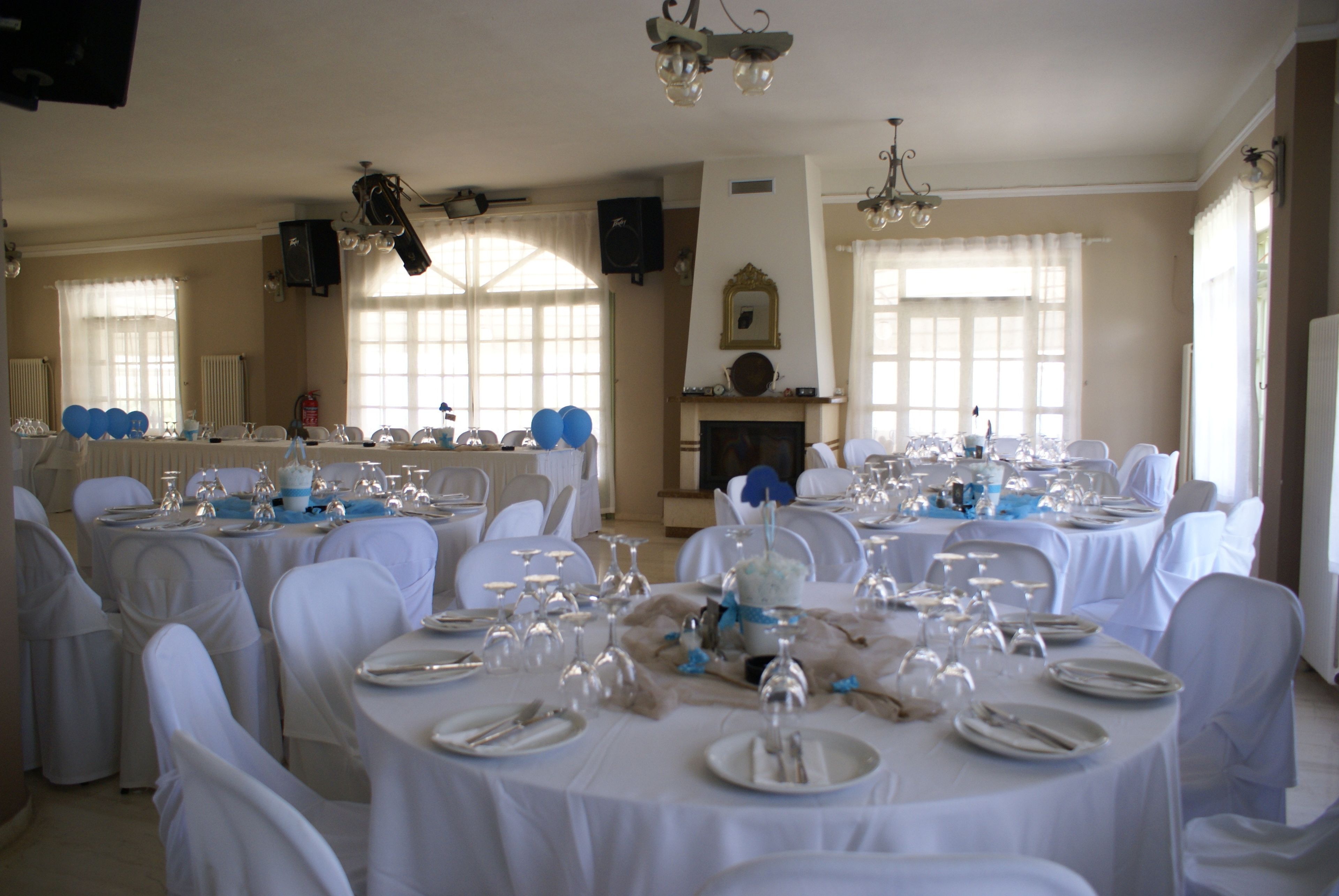 Un salón de banquetes con mesas redondas dispuestas para un evento formal, con manteles y sillas blancas, una chimenea y ventanas amplias.