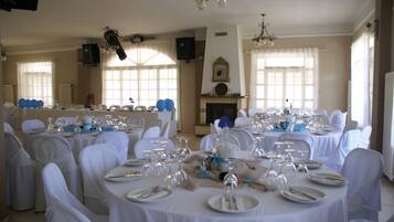Un salón de banquetes con mesas redondas dispuestas para un evento formal, con manteles y sillas blancas, una chimenea y ventanas amplias.