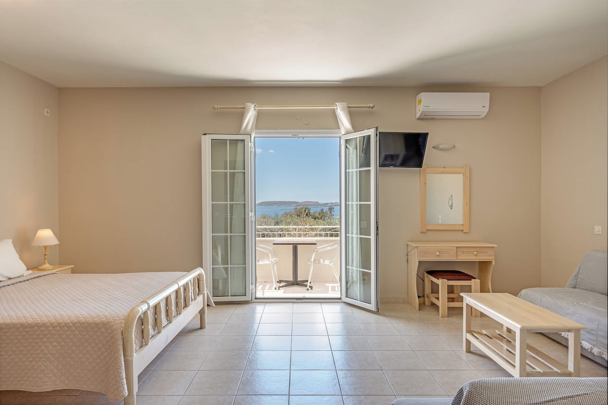 A bedroom with a bed, a desk, a chair, a small table, and a balcony with a view.