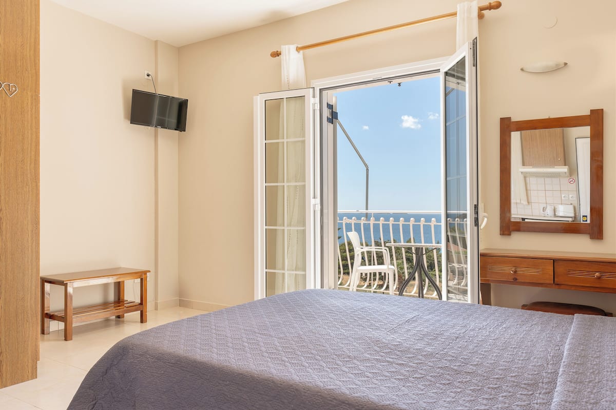 A hotel room with a bed, a TV mounted on the wall, a wooden desk, and a balcony with a view of the sea.