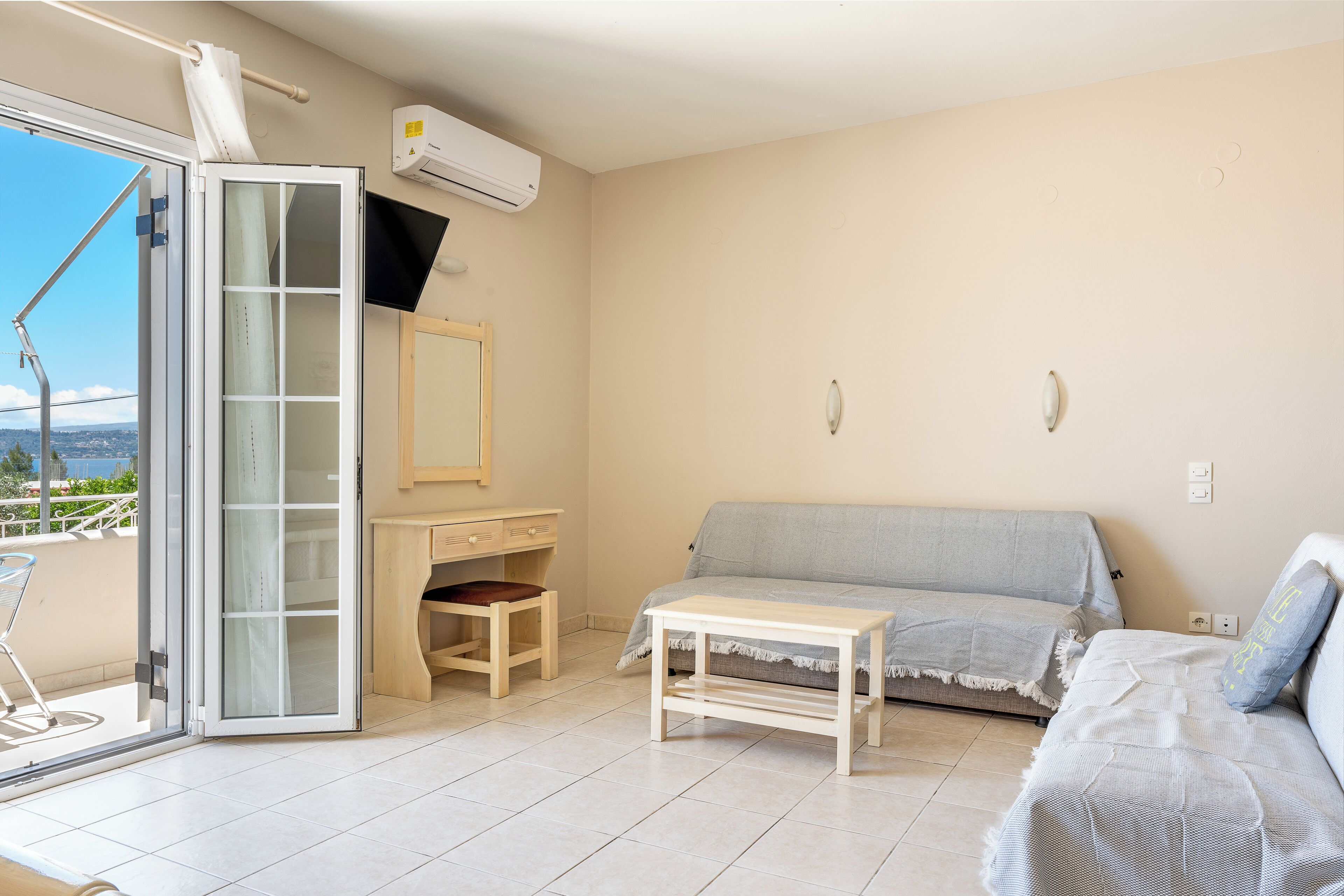A room with a sliding glass door leading to a balcony, a sofa, a small table, a desk with a chair, and a TV mounted on the wall.