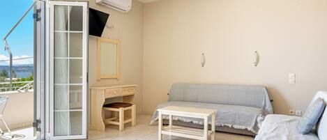 A room with a sliding glass door leading to a balcony, a sofa, a small table, a desk with a chair, and a TV mounted on the wall.