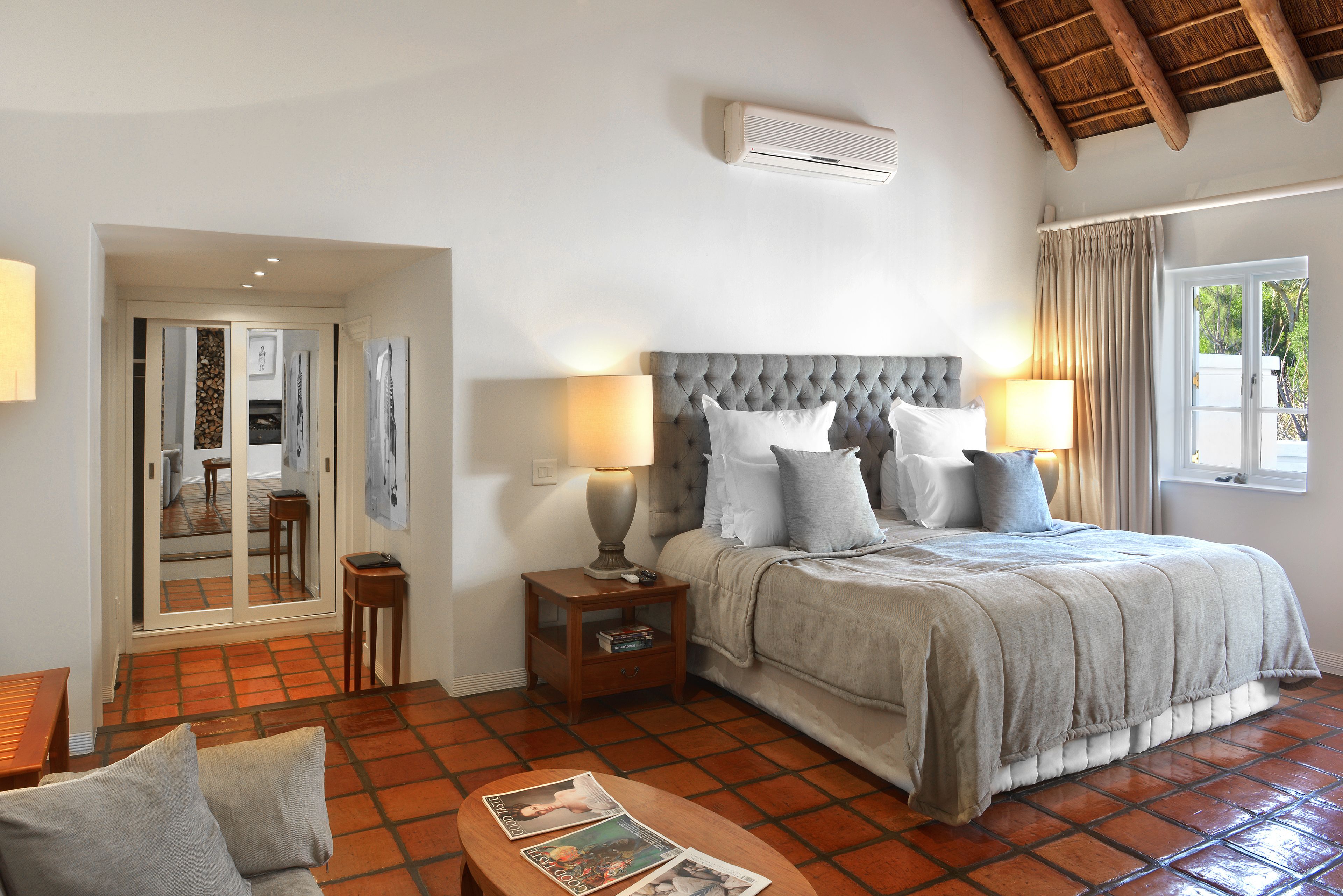 A bedroom with a large bed, nightstands, a wooden table, and a view of the outside.