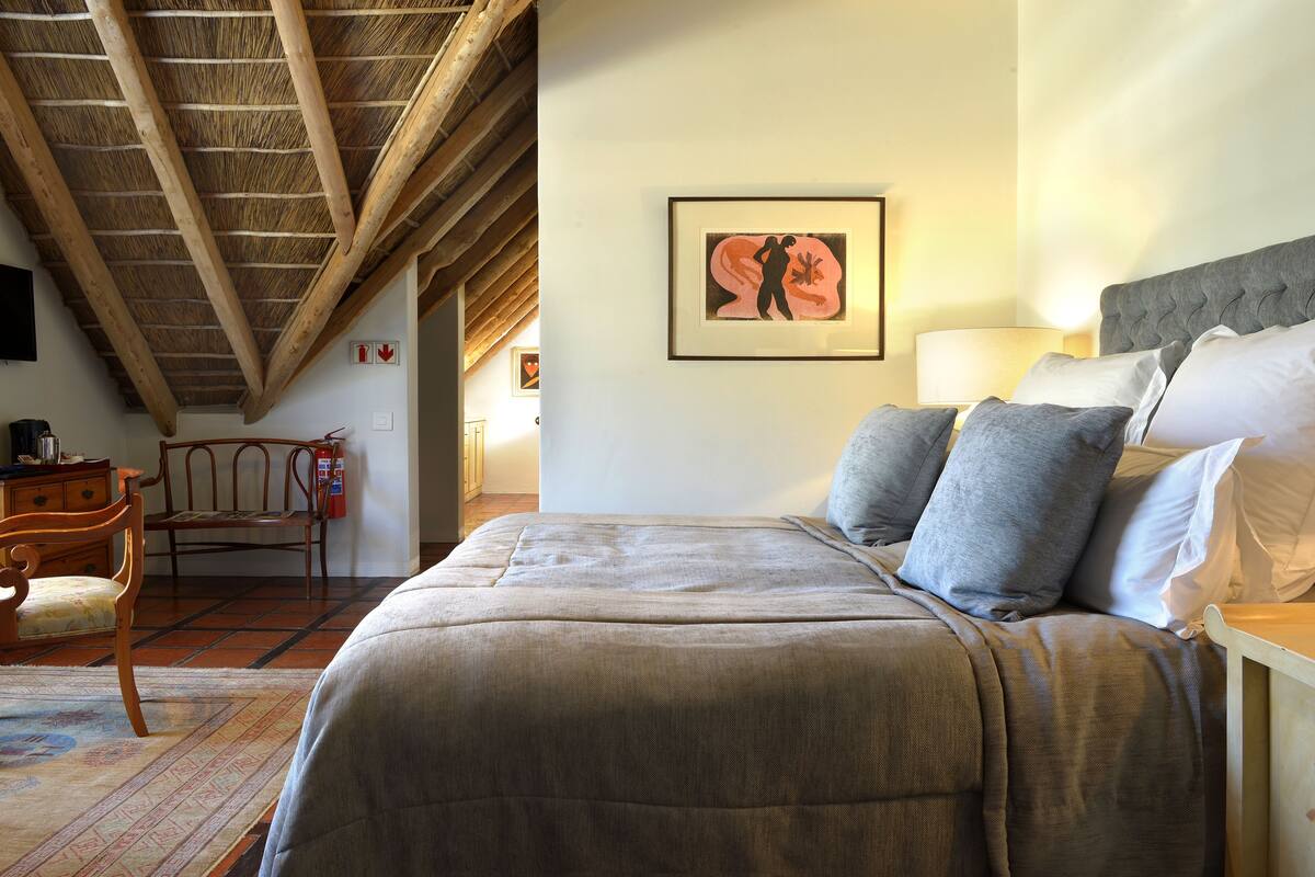Een slaapkamer met een houten plafond, een bed met grijs en wit beddengoed, een kunstwerk in een lijst aan de muur, en een stoel in de hoek.