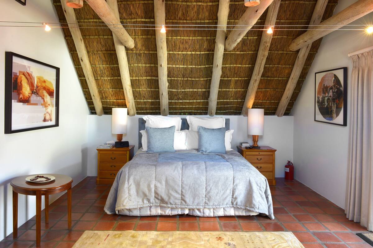 Een slaapkamer met rieten dak, een bed met grijze en witte dekbedovertrek, twee nachtkastjes met lampen, en een klein rond tafeltje met een bord eten.
