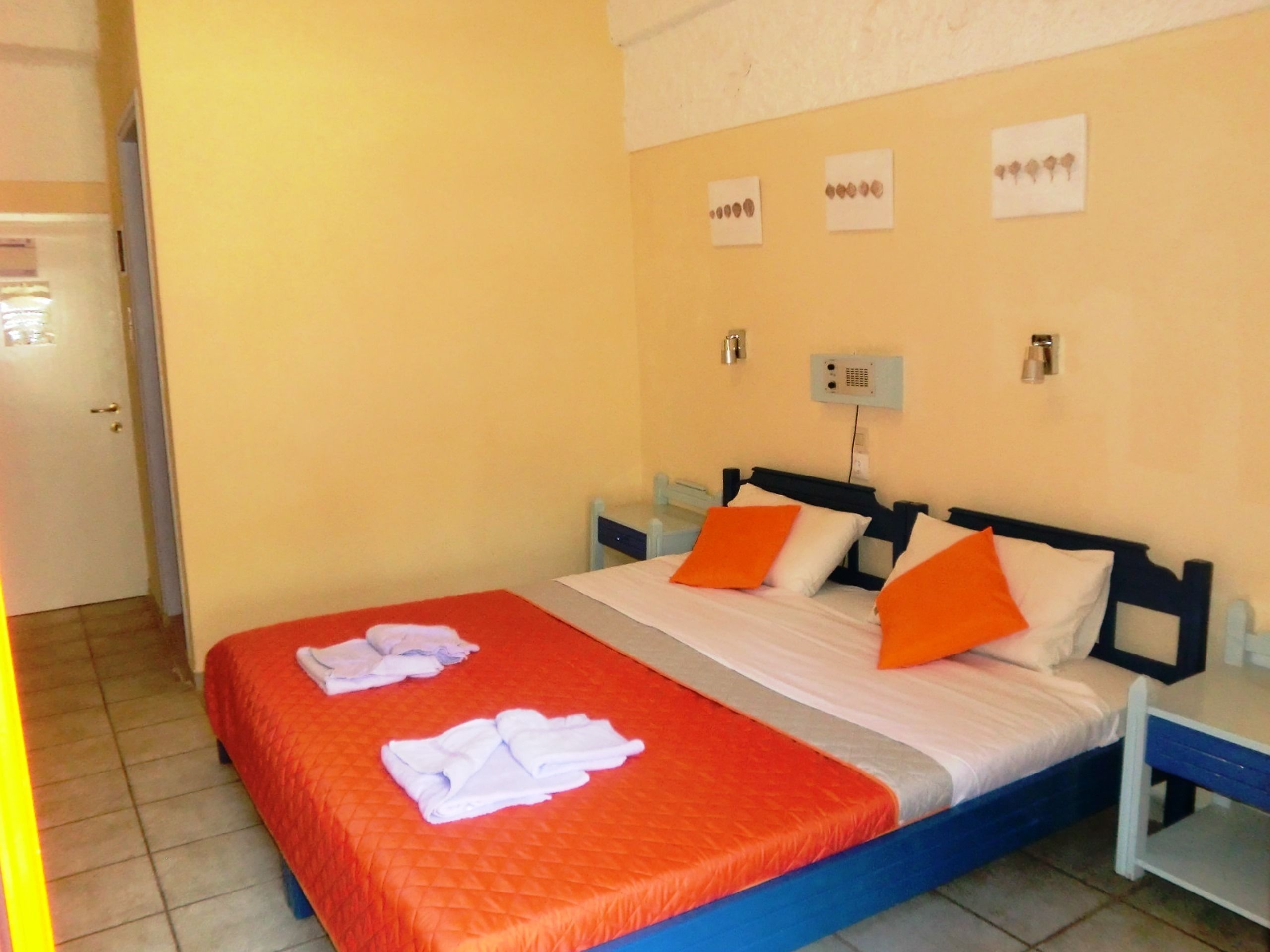 A hotel room with two beds, orange bedding, white towels, and a simple wall-mounted light fixture.