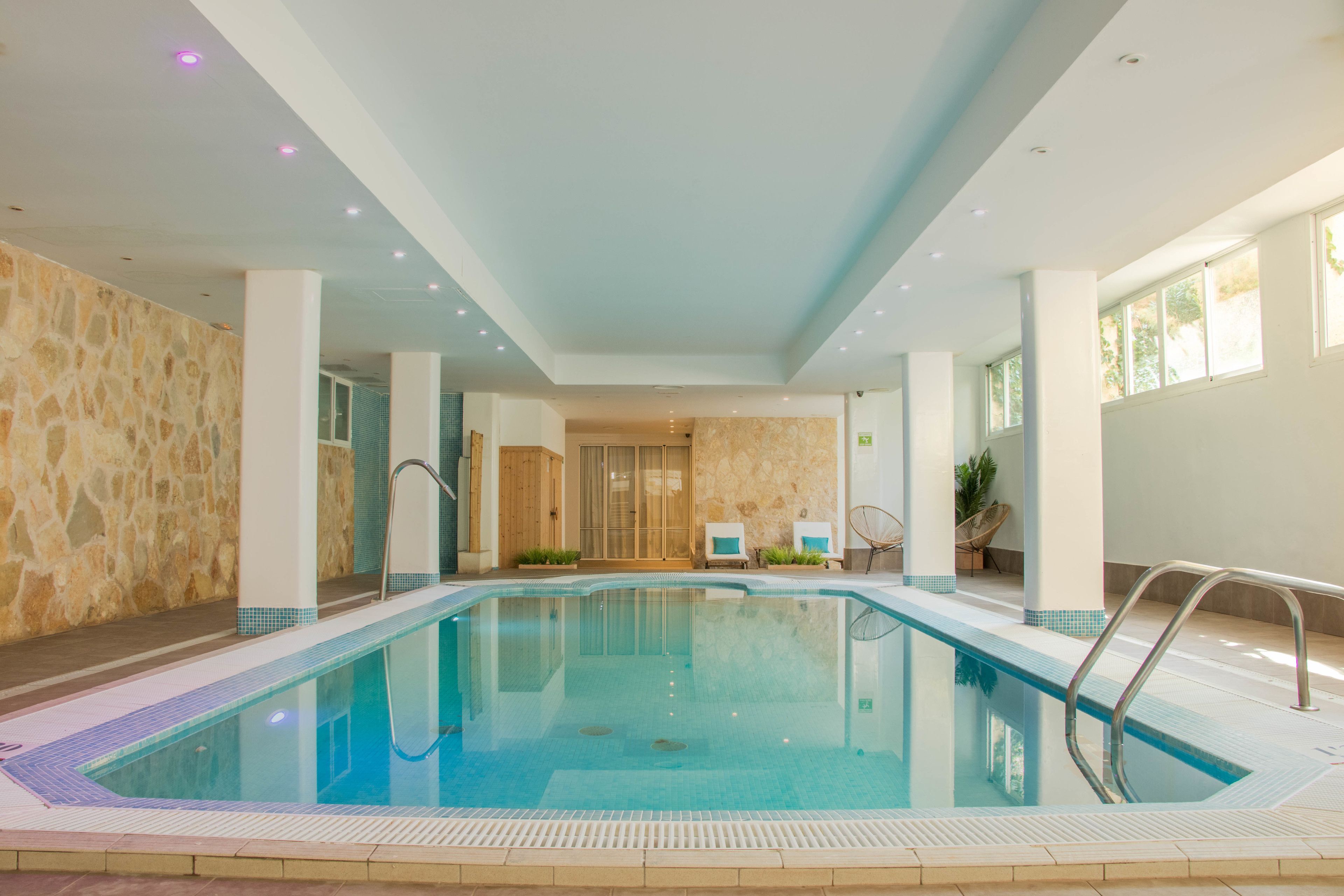 An indoor swimming pool with clear water, surrounded by stone walls and columns, and a view of a wooden door in the background.